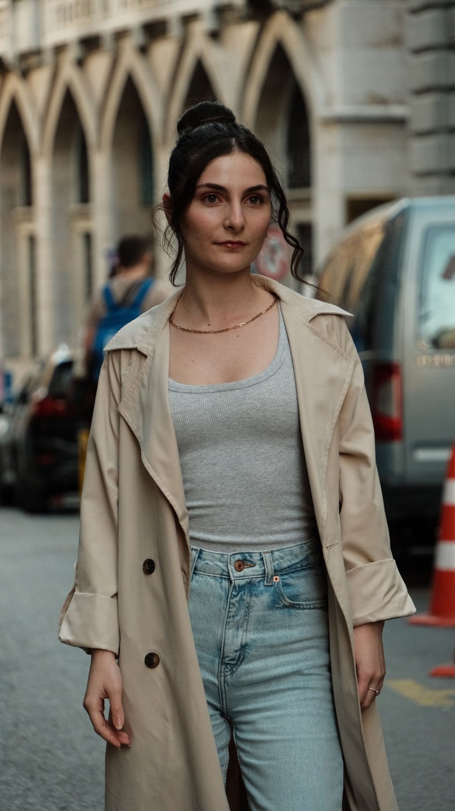 Open beige double-breasted trench coat over a grey ribbed tank top and light-blue jeans, with a fine necklace; cropped above the feet.