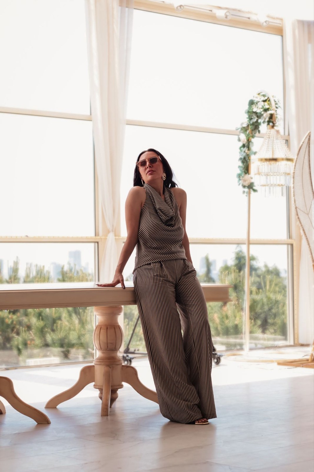 Taupe sleeveless draped top and matching wide-leg trousers with sunglasses; standing by a window.
