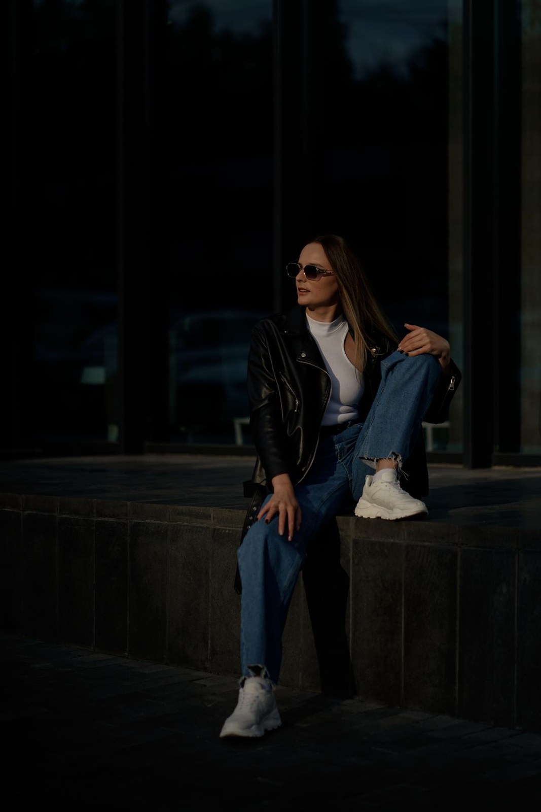 Blue denim jacket and jeans, white top and white trainers, seated in the city.