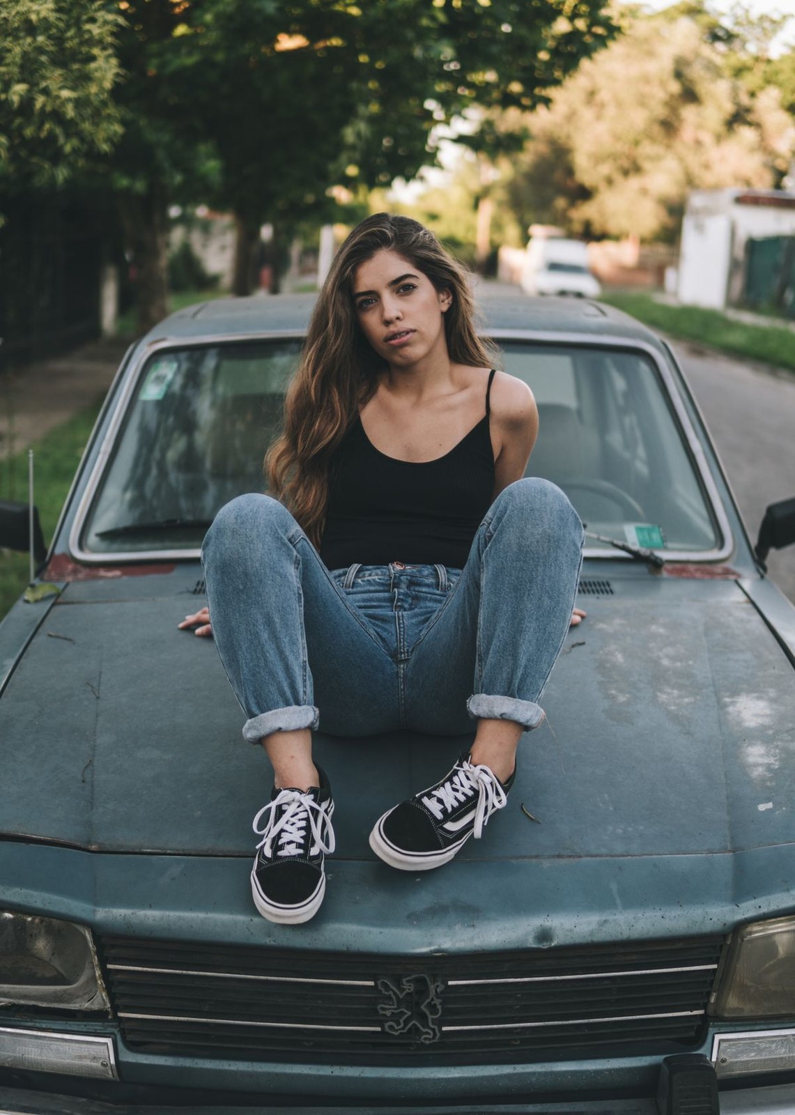 Black tank, cuffed blue jeans and black-and-white low-top trainers, seated on a car.