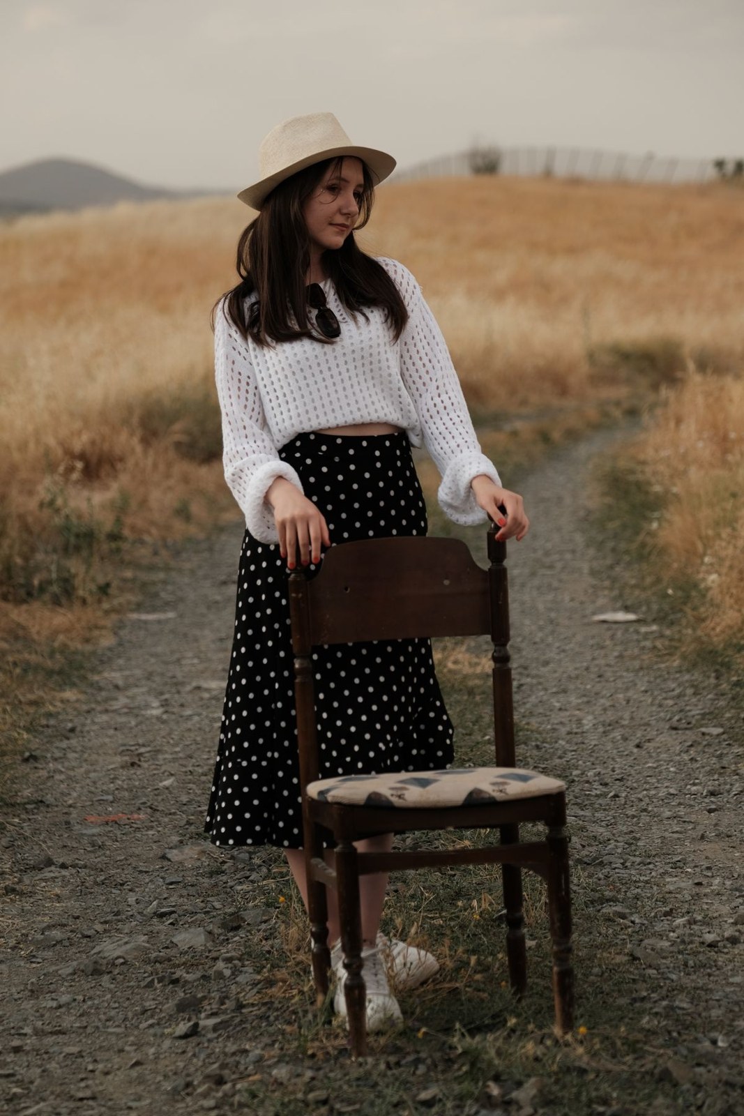 White open-knit sweater, long black skirt with small white dots, pale wide-brim hat and brown bag.