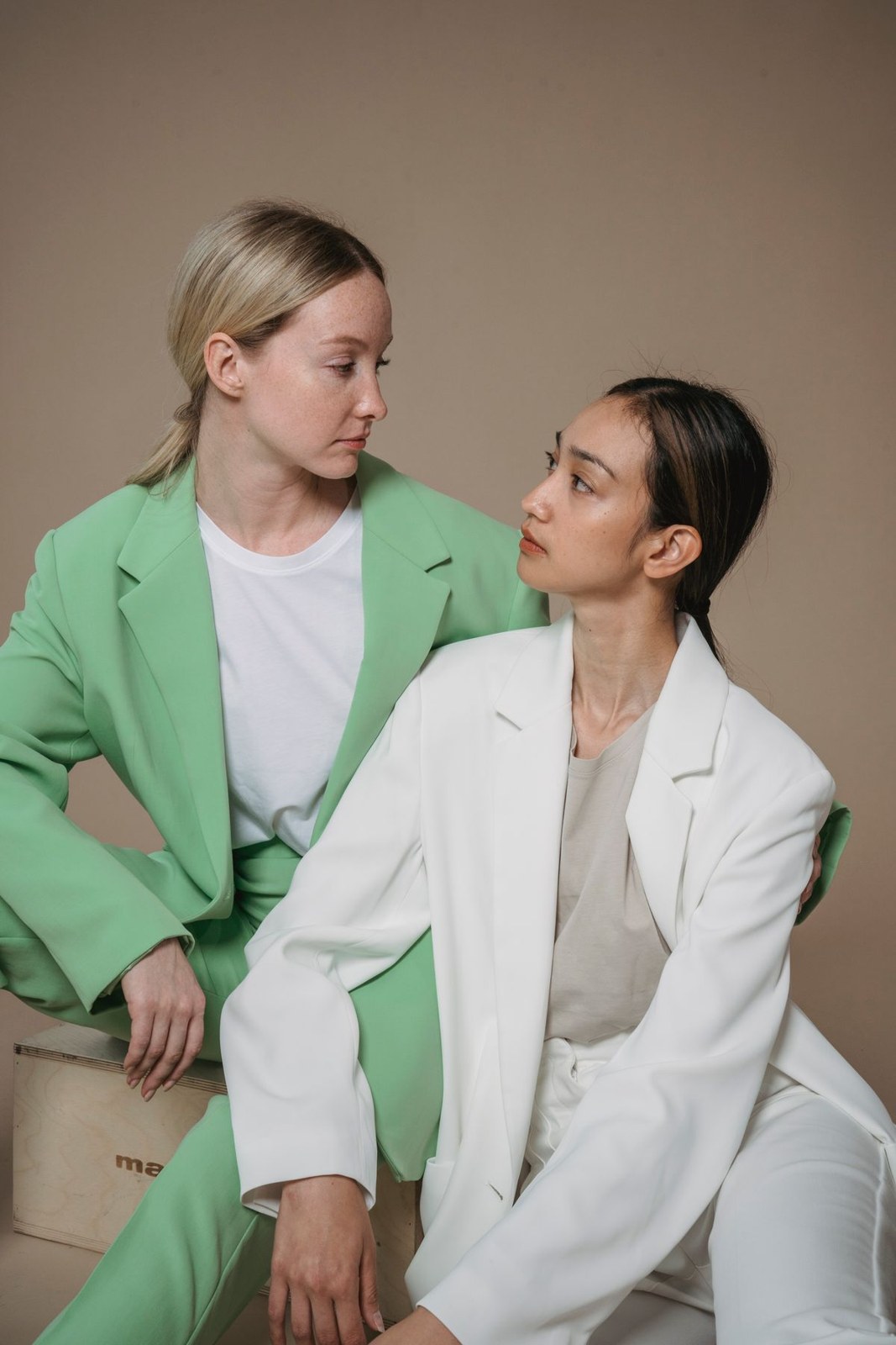 Seated women in a green trouser suit with white T-shirt and white suit with grey top.
