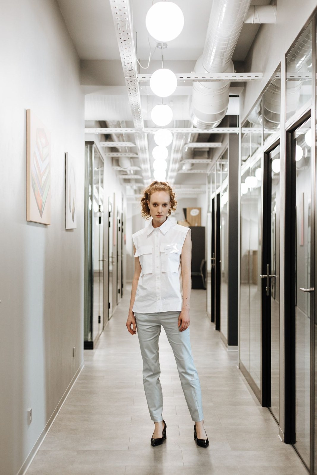 White sleeveless collared blouse, pale-grey ankle trousers and dark pumps in an office corridor.