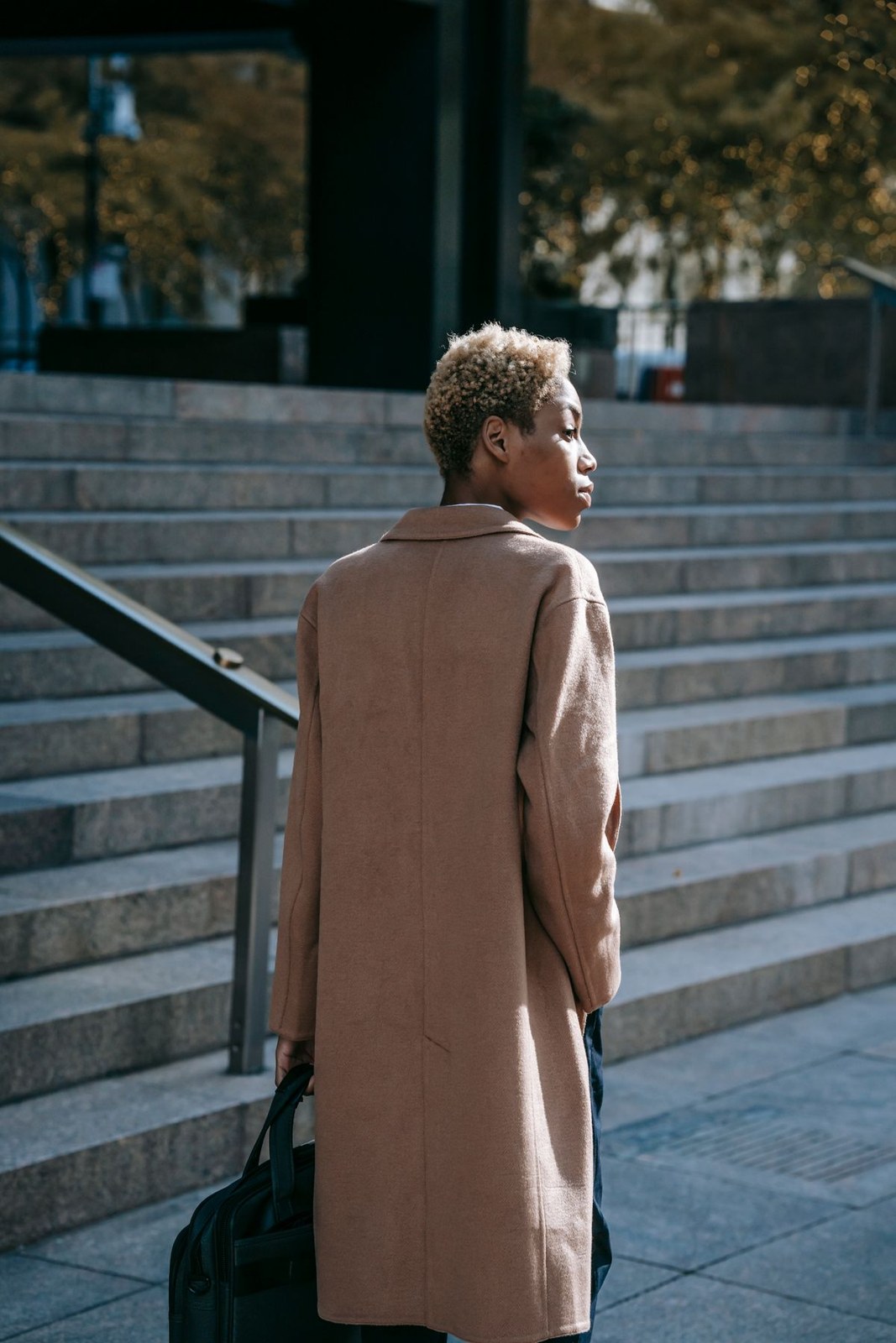 Long camel coat viewed from behind, with dark trousers and a dark handbag; lower legs cropped.