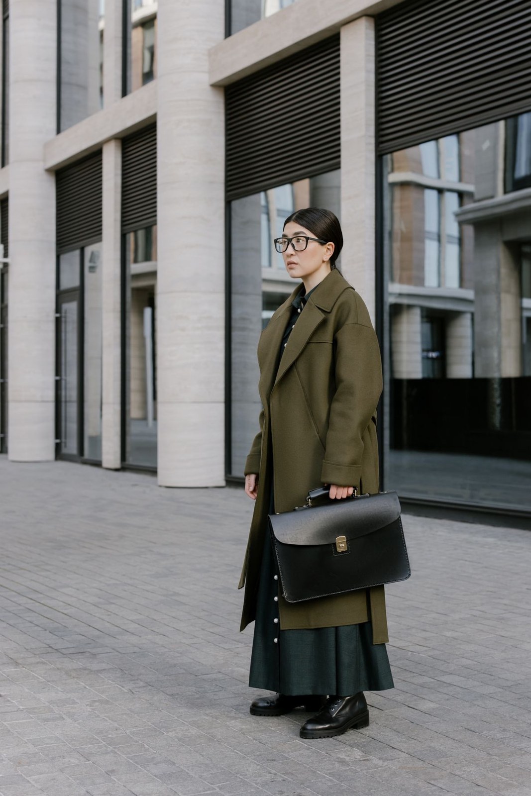 Olive double-breasted coat over a long dark-green skirt, dark boots, glasses and a black structured bag.