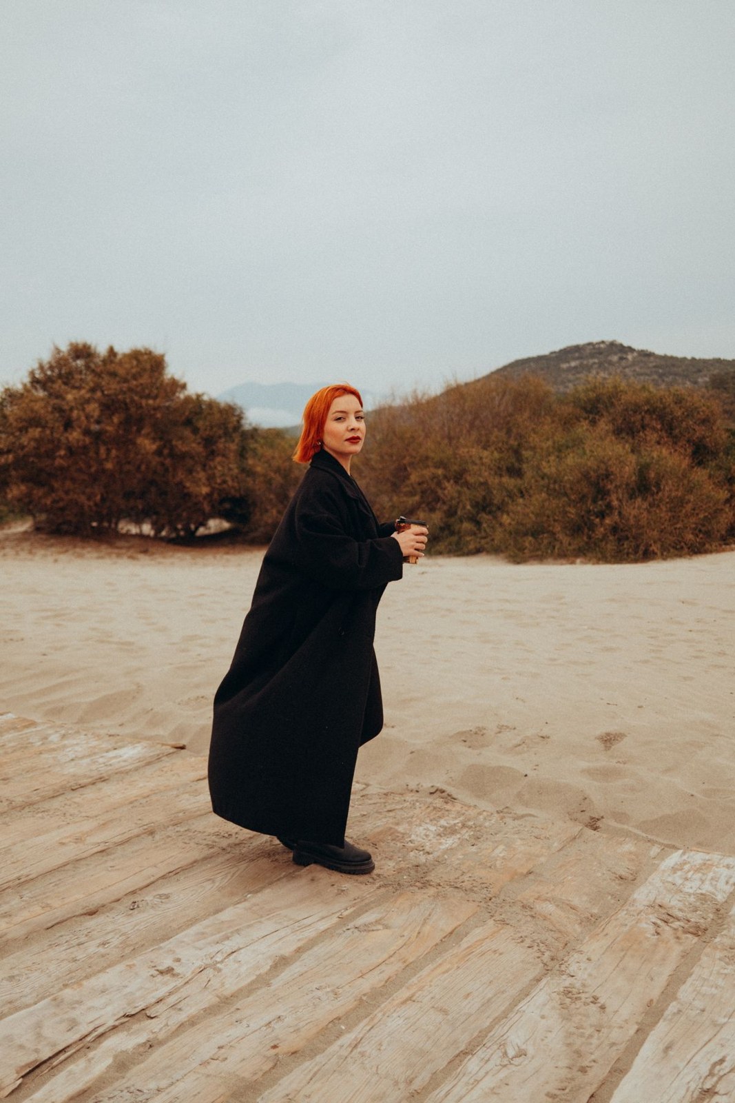 Long black coat and dark footwear in a windy outdoor sandy setting.