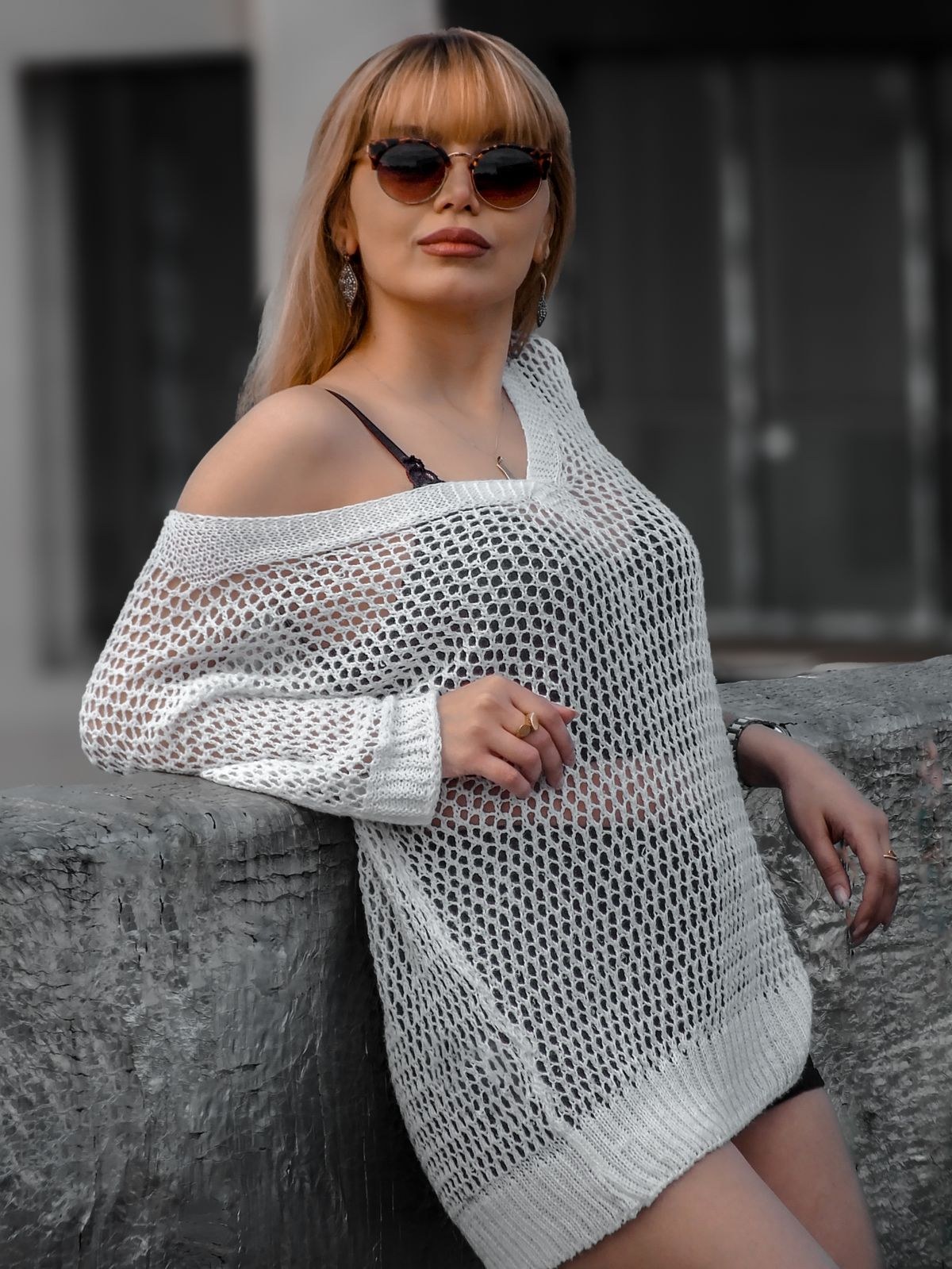 White open-knit sweater worn off one shoulder over a black strappy top, with sunglasses.