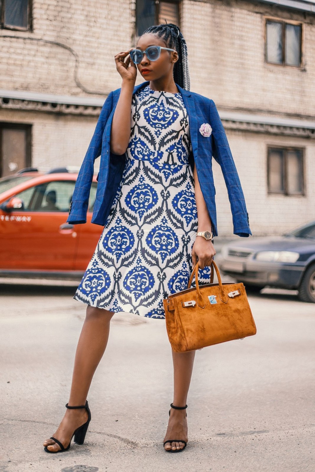 Blue-and-white printed sleeveless dress, blue blazer over the shoulders, tan structured bag and black strappy block heels.