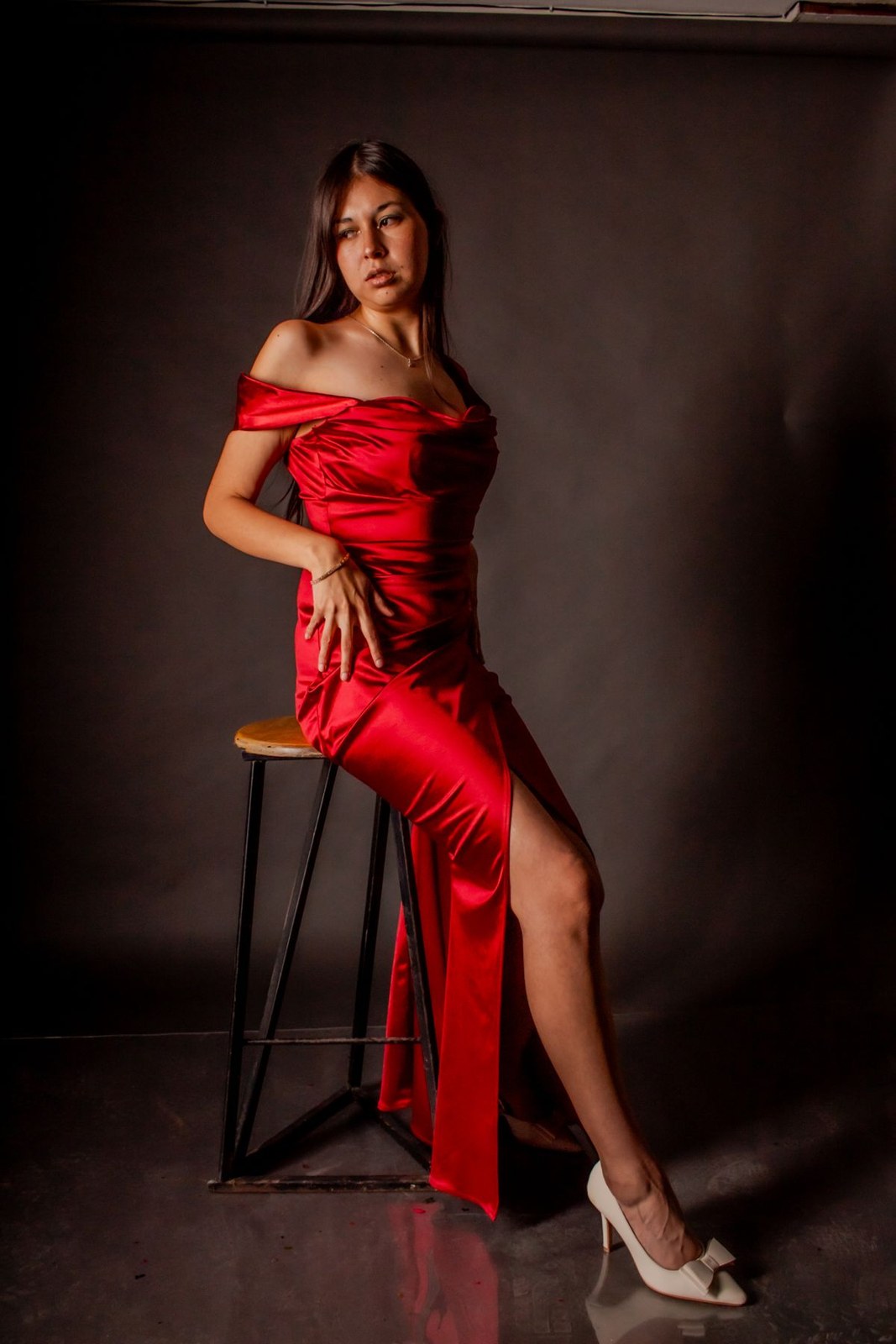 Red off-shoulder long dress with a thigh slit and pale heels, seated on a studio stool.