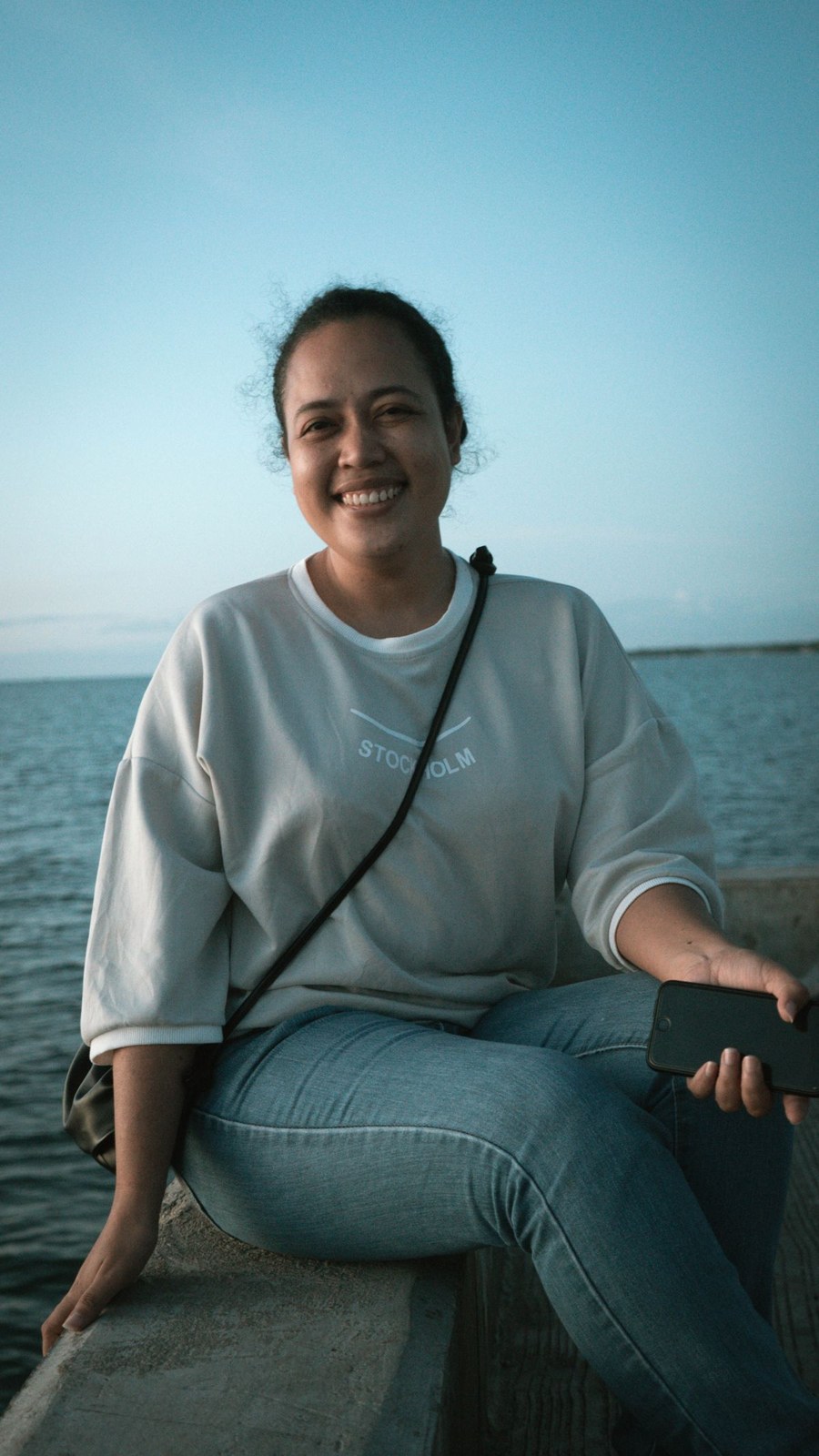 Hellgraues weites Sweatshirt, blaue Jeans mit Umschlag und kleine schwarze Umhängetasche, sitzend am Meer.